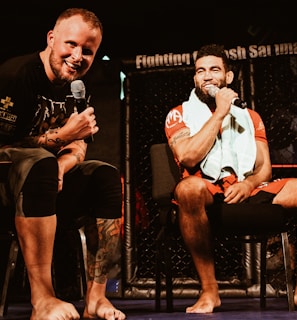 Two individuals are seated on a stage, each holding a microphone and smiling. They are in a sports-related setting with one wearing a casual black outfit and the other in sportswear, with a towel around his neck. The background includes elements of a fighting or martial arts cage.
