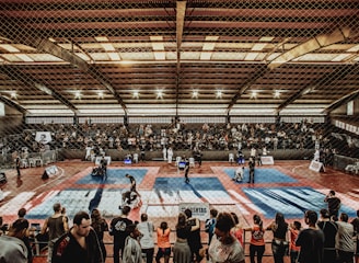 A large indoor arena with a high ceiling, filled with an audience seated on bleachers. In the center, two fighters are engaged in a martial arts competition on blue and red mats. The scene is captured through a mesh fence, adding a textured foreground.