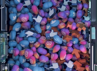 A top-down view of a vibrant pile of colorful icons and symbols representing various social media platforms. Icons include blue Twitter birds, Instagram logos, Facebook-like thumbs up, and orange up arrows. The symbols are situated inside a futuristic, mechanical enclosure, possibly resembling a server or machine.