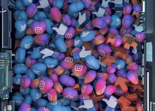 A top-down view of a vibrant pile of colorful icons and symbols representing various social media platforms. Icons include blue Twitter birds, Instagram logos, Facebook-like thumbs up, and orange up arrows. The symbols are situated inside a futuristic, mechanical enclosure, possibly resembling a server or machine.