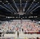 An indoor sports arena filled with rows of tables, each occupied by people engaged in competitive activities. The ceiling is high, with numerous lights illuminating the large space. The audience seating on either side is empty, and there are flags hanging from the upper level.