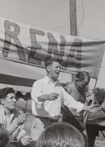 A group of men, some shirtless, are gathered around a platform or stage. A man in a white shirt appears to be speaking or gesturing to a crowd. A large sign with the word 'ARENA' is prominently displayed in the background. The scene seems to be set outdoors on a sunny day.