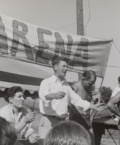 A group of men, some shirtless, are gathered around a platform or stage. A man in a white shirt appears to be speaking or gesturing to a crowd. A large sign with the word 'ARENA' is prominently displayed in the background. The scene seems to be set outdoors on a sunny day.
