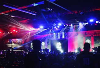 A dimly lit indoor arena filled with an audience. Bright stage lights in red and blue illuminate multiple large screens displaying graphics.