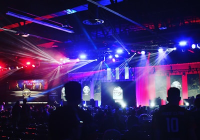 A dimly lit indoor arena filled with an audience. Bright stage lights in red and blue illuminate multiple large screens displaying graphics.