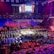 An indoor arena is filled with people watching a wrestling match. The ring is centrally located, and surrounded by audience members sitting in tiered seating. The arena is illuminated with red and blue lights, and a large overhead screen displays 'Sunday Stunner.' Spectators are engrossed in the match taking place in the well-lit ring.