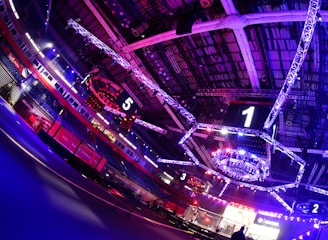An indoor arena with vibrant purple and blue lighting, featuring large numbered screens suspended from the ceiling. The venue has a high ceiling with intricate metal truss structures and hanging lights, creating a futuristic and energetic atmosphere.