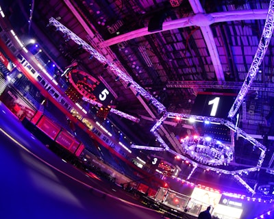 An indoor arena with vibrant purple and blue lighting, featuring large numbered screens suspended from the ceiling. The venue has a high ceiling with intricate metal truss structures and hanging lights, creating a futuristic and energetic atmosphere.
