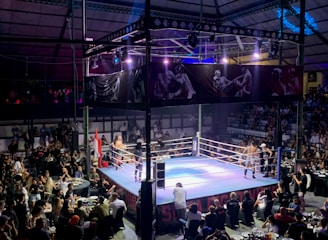 A crowded indoor arena with a boxing ring at the center, surrounded by an audience seated at tables. The scene is illuminated by overhead lights, casting a vibrant glow over the ring where two fighters are preparing for a match. Spectators are engaged, capturing the event with cameras and enjoying the lively atmosphere.