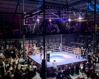 A crowded indoor arena with a boxing ring at the center, surrounded by an audience seated at tables. The scene is illuminated by overhead lights, casting a vibrant glow over the ring where two fighters are preparing for a match. Spectators are engaged, capturing the event with cameras and enjoying the lively atmosphere.