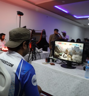 A group of people is gathered in a room watching and participating in a video game competition. Players and spectators are focused on the monitors displaying a fighting game. The setting includes tables with equipment and chairs arranged around the venue, with a noticeable intensity in the participants’ engagement.