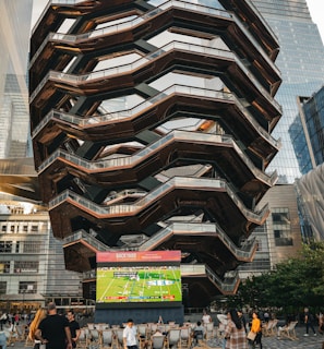 A large, spiral architectural structure with multiple levels stands prominently in an urban setting. In front of the structure, a large screen displays a sports broadcast, and several people are gathered around, some sitting and others standing. The scene is set outdoors, with towering modern buildings surrounding the area. The ground is paved with a geometric pattern of gray and white tiles.